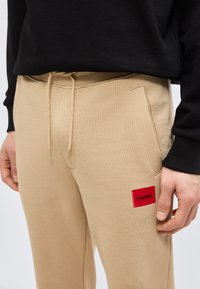 Μπεζ παντελόνι φόρμας με πλαϊνές τσέπες και κορδόνι στη μέση, με κόκκινο κέντημα λογότυπου HUGO στον μηρό, συνδυασμένο με μαύρο μακρυμάνικο τοπ.