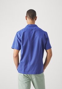 Kortärmad skjorta i djupblå bomull, med krage och ryggdetalj. Bärs med ljusgröna byxor, vilket ger en avslappnad look.