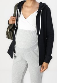 Sweat à capuche noir zippé avec cordons blancs sur un haut croisé blanc, assorti à un pantalon de jogging gris à taille élastique. Tissu lisse, coupe décontractée.