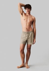 Costume da bagno beige con cintura elastica e dettaglio con stringa, realizzato in tessuto morbido, indossato da un modello a piedi nudi.