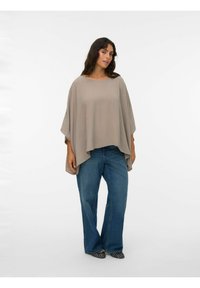 Beige oversized poncho med et afslappet, flydende design. Har en rund halsudskæring og brede ærmer, som er kombineret med blå trompetbukser og sorte sko.