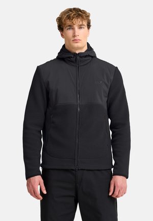Junger Mann mit lockigen Haaren, der eine schwarze Fleecejacke mit Reißverschluss und Kapuze trägt und vor einem einfarbigen hellen Hintergrund steht.
