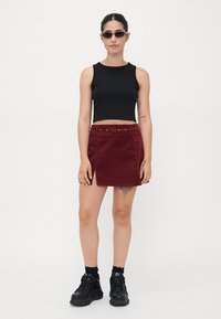 Dieprode corduroy rok met een riem in de taille en knoopdetails, gecombineerd met een zwarte crop top en zwarte chunky sneakers. Rechthoekige zonnebril gedragen.