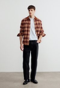 Νέος άνδρας στέκεται με το ένα χέρι στην τσέπη, φορώντας ένα καφέ καρό πουκάμισο πάνω από λευκή μπλούζα, μαύρο παντελόνι και μαύρα παπούτσια μπροστά σε απλή λευκή τοίχο.