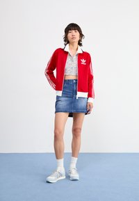 Červená športová bunda s bielymi pruhmi a golierom, kvetinový crop top, džínová sukňa a svetlomodré tenisky s bielymi ponožkami. Svetlomodré pozadie.