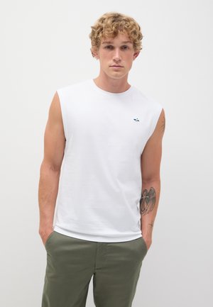Camiseta sin mangas blanca hecha de una tela suave, con un pequeño logo azul y verde en el pecho. Combinada con pantalones verde oliva.