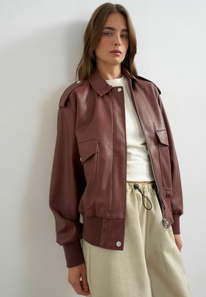 Kvinde iført en brun læderjakke over en hvid top og beige bukser med snøre, stående mod en ensfarvet lys væg.