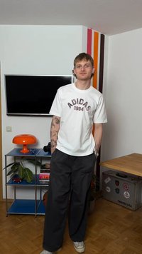 Νέος άνδρας με τατουάζ, ντυμένος με λευκό μπλουζάκι Adidas και μαύρο παντελόνι, στέκεται σε δωμάτιο με τηλεόραση, πορτοκαλί φωτιστικό, ράφια και διακόσμηση τοίχου με ρίγες.