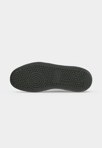 Semelle de chaussure en caoutchouc noir avec des motifs de crampons circulaires et linéaires et un patch logo rectangulaire au centre.