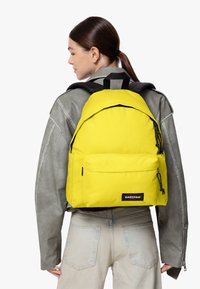 Mochila amarilla con un tejido suave y duradero y un bolsillo frontal. Presenta detalles en negro y correas ajustables. Llevada en la espalda de una persona.