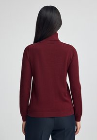 Frau mit langen dunklen Haaren, die einen burgunderfarbenen Rollkragenpullover und dunkle Hosen trägt, von hinten vor einem schlichten Hintergrund gezeigt.