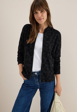 Schwarze Bomberjacke mit Leopardenmuster, Reißverschluss vorne, gerippte Bündchen und glatte Stoffoberfläche, kombiniert mit blauen Jeans und einer beigen Tasche.