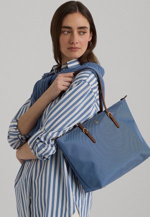 Vrouw met bruin haar, gekleed in een blauw-wit gestreept shirt, met een grote blauwe handtas over de schouder tegen een effen achtergrond.