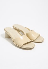 Beige open-toe mule sandalen met lage blokhakken en brede voorbanden, gepresenteerd op een witte achtergrond.