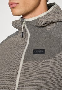 Siva zip-up dukserica s teksturiranom, uzorkovanom tkaninom. Ima kapuljaču, kontrastni bijeli zatvarač i crnu oznaku s logotipom na prsima.