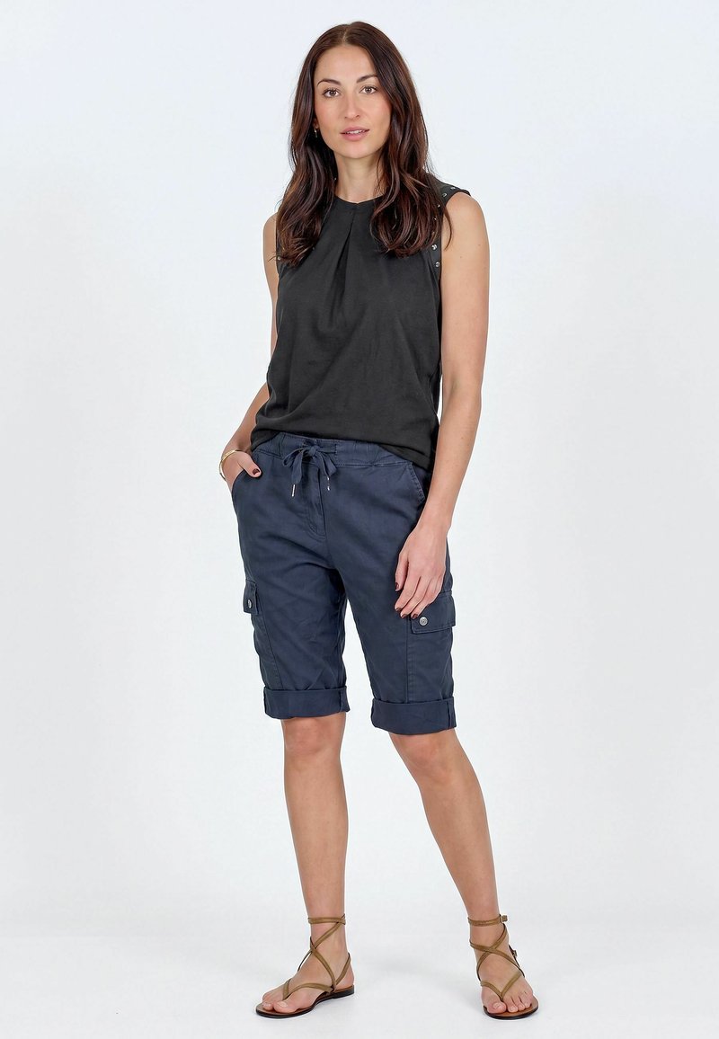 Femme debout portant un haut noir sans manches, un short cargo bleu marine roulé à l'ourlet, et des sandales plates marron à brides.