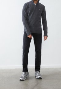 Dunkelgrauer Pullover mit Reißverschluss bis zur Brust, gerippten Bündchen und Saum, getragen mit schwarzer Jeans und silbernen Sportschuhen.