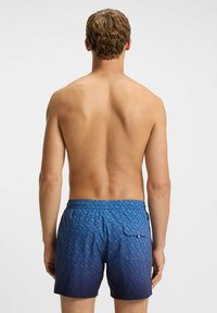 Male with short curly hair shown from the back wearing blue patterned swim shorts with a back pocket on a white background.
