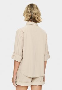 Beige, lightweight, collared shirt with long sleeves rolled up, featuring a rounded hemline and a relaxed design.