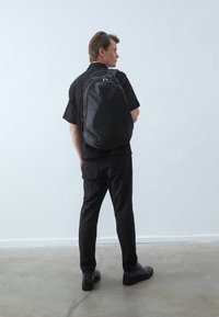 Svart ryggsekk med teksturert finish, utstyrt med justerbare stropper, glidelås og en slank, strømlinjeformet design. Båret av en modell.