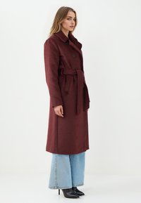 Lungo cappotto bordeaux con vita cinturata, chiusura a bottoni e ampio colletto. Indossato sopra jeans a vita alta blu chiaro e tacchi neri.