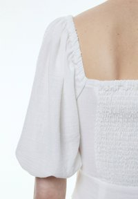 Weiße Bluse mit puffärmel und einer gerafften Rückendetail. Der Stoff wirkt leicht und hat eine weiche Textur.