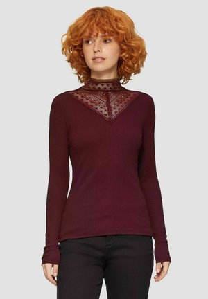 Bordeauxrode lange mouwen shirt met een hoge halslijn en kantdetails in een V-vorm. Geribbelde textuur en aansluitend ontwerp, gedragen met zwarte broek.