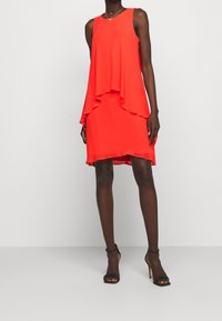 Kvinna i ärmlös klänning i ljus röd färg med lager och svarta högklackade sandaler, som står mot en enkel ljus bakgrund.