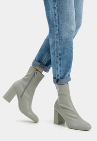 Zielone botki z sztucznej skóry za kostkę na słupkowym obcasie z bocznym zamkiem; para zawiniętych niebieskich dżinsów widoczna nad botkami.