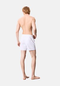 Costumi da bagno bianchi con fascia elastica in vita. Vista da dietro, che mostra un design semplice e una lunghezza fino al ginocchio. Nessun motivo o dettaglio presente.