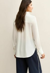 Blusa blanca de manga larga con cuello puntiagudo y puños con botones; textura suave. Combinada con pantalones azul marino a rayas finas.