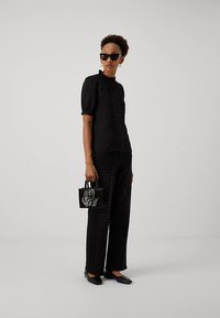 Woman wearing black sunglasses, black short-sleeve blouse, black wide-leg pants with subtle dots, holding a black handbag with a large flower detail.