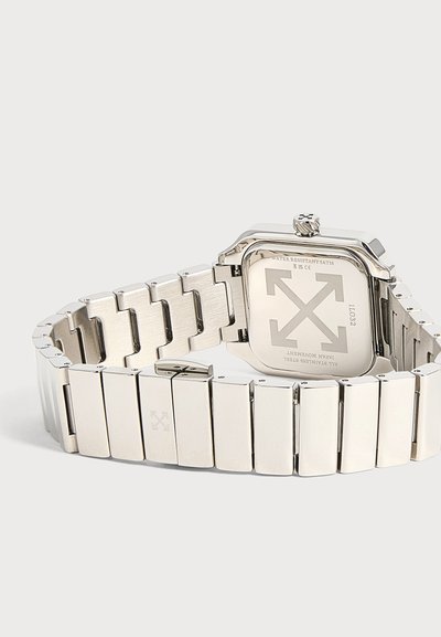 Montre-bracelet en métal argenté avec un fond de boîtier carré indiquant la résistance à l'eau et le logo de la marque composé de quatre flèches.