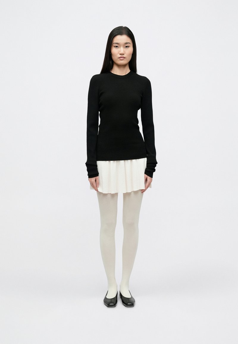 Pulover negru cu mâneci lungi, cu model rib, asortat cu o fustă mini plisată albă. Modelul poartă ciorapi albi transparenți și pantofi negri fără toc.