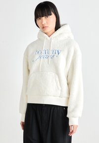Cream-colored fleece hoodie with drawstrings, embroidered "Tommy Jeans" logo in blue, kangaroo pocket, and ribbed bottom hem.