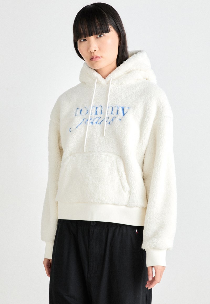 Cream-colored fleece hoodie with drawstrings, embroidered "Tommy Jeans" logo in blue, kangaroo pocket, and ribbed bottom hem.