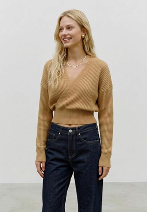 Femme souriante aux longs cheveux blonds porte un pull cache-cœur côtelé couleur camel et un jean taille haute bleu foncé devant un fond clair uni.