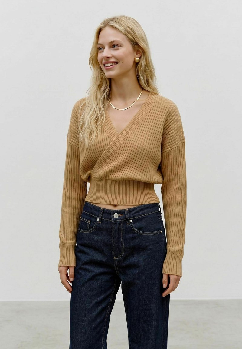 Femme souriante aux longs cheveux blonds porte un pull cache-cœur côtelé couleur camel et un jean taille haute bleu foncé devant un fond clair uni.