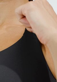 Czarny sportowy top z gładkiego, elastycznego materiału z okrągłym dekoltem i minimalną ilością szwów, podkreślony ręką trzymającą materiał.