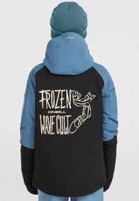 Zwart en blauwe jas met capuchon, voorzien van een grote grafische print op de achterkant met de tekst “FROZEN WAVE CULT” in crème. Gestructureerd materiaal.