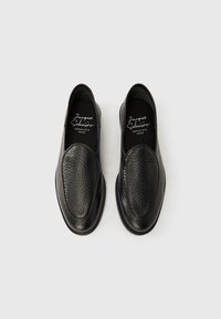 Chaussures noires à enfiler avec dessus en cuir texturé, bout ovale et semelle lisse. Logo visible sur la semelle intérieure. Design classique et minimaliste.