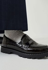 Mocassin noir brillant texturé avec semelle épaisse, porté avec des chaussettes côtelées gris clair et un pantalon à fines rayures foncé.