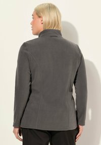 Personne blonde de dos portant une veste en polaire grise foncée ajustée à manches longues et un pantalon noir, devant un fond clair.