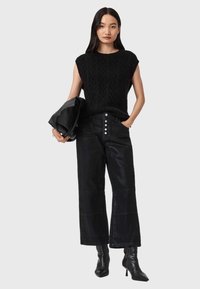 Top senza maniche in maglia nera, abbinato a jeans neri a vita alta con chiusura a bottoni. Il modello tiene una borsa nera, accentuata da stivali a punta.