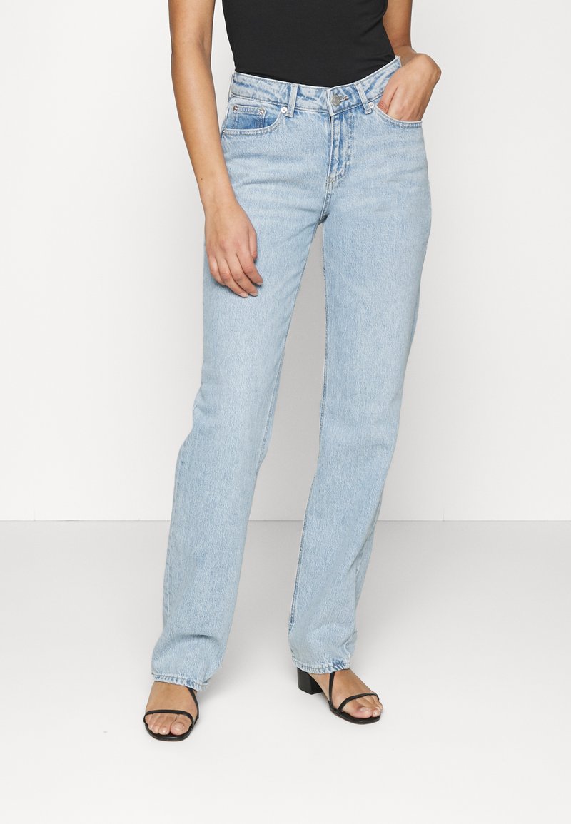 Svetlo modri jeans z ravnim krojem, visokim pasom in klasičnim petim žepom. Obut z črno majico in črnimi sandalmi na peto.