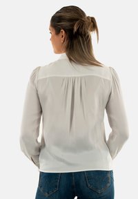 Blouse blanche à manches longues avec un dos plissé, texture douce et poignets à boutons, portée avec un jean bleu présentant un motif de couture distinct.