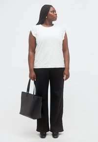 Γυναίκα με μακριά πλεξούδα, φορώντας λευκό τοπ με σχέδιο τρυπών και μαύρο παντελόνι, κρατώντας μια μαύρη τσάντα, κοιτάζει προς τα πλάγια.