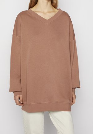 Femme portant un sweat-shirt marron à col en V oversize et un pantalon blanc, debout devant un fond clair uni.