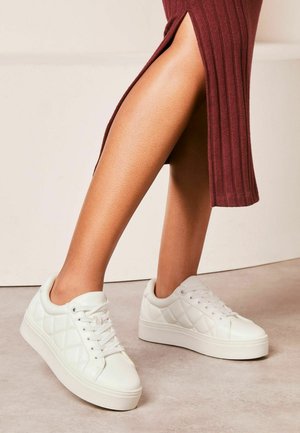 White, textured sneakers with quilted pattern and flat sole. Laces in front, rounded toe, paired with a ribbed burgundy dress with a side slit.