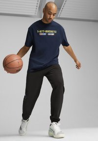 Tumši zila t-krekls ar tekstu "1-877-BUCKETZ", melnas sporta bikses un baltas kedas ar zaļiem akcentiem, driblē basketbola bumbu telpās.